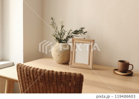 Elegant artistic mockup template. Black vertical photo frame on wooden table desk. Vase with olive tree branches, cup of tea, coffee. Poster display. Blurred rattan chair. Summer Mediterranean design. 119486603