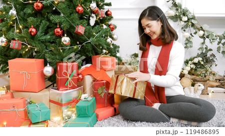 Close up woman hands wrap present gift in cozy living room. Woman wrapping christmas gift box under xmas tree. Asian woman decor bauble ball christmas tree in xmas holiday winter seasonal celebration 119486895