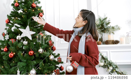 Woman hands decorate Christmas tree red ball, bauble, snow flake. Asian woman decor bauble ball christmas tree in xmas holiday winter seasonal celebration. Close up hands holding silver star decorate 119486898