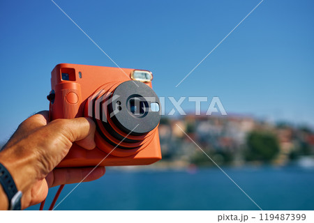 Close-up of tourist hand holding an orange instant camera in retro style with coastal town and blue sea in background. Summer travel concept. Equipment for capturing moments Close-up of tourist hand holding an orange instant camera in retro style with coastal town and blue sea in background. Summer travel concept. Equipment for capturing moments 119487399