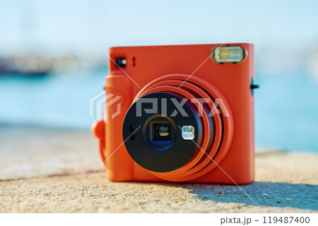 Vintage orange instant camera on rock by seaside with blurred coastal town in background. Summer travel concept. Equipment for capturing moments Vintage orange instant camera on rock by seaside with blurred coastal town in background. Summer travel concept. Equipment for capturing moments 119487400