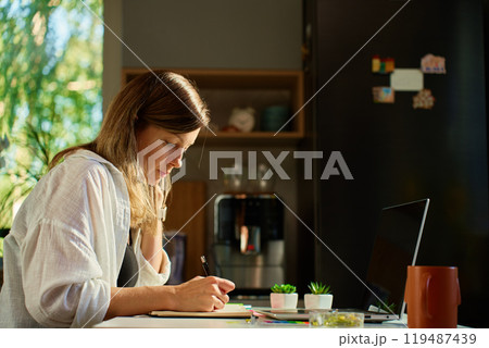Freelance woman sitting at kitchen table using laptop and writing in notebook. Remote work at home office 119487439