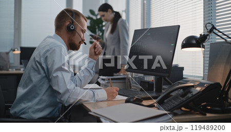 In modern office: Financial trader in headset works on computer, communicates with client 119489200
