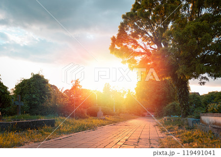 The path leading straight through the Christian cemetery in the rays of the setting sun. Sunset in background. A secluded atmosphere The path leading straight through the Christian cemetery in the rays of the setting sun. Sunset in background. A secluded atmosphere 119491041