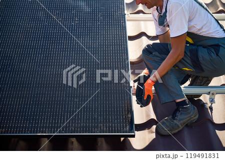 Worker building photovoltaic solar panel system on rooftop of house. Man engineer in gloves installing solar module with help of hex key outdoors. Alternative and renewable energy generation concept. 119491831