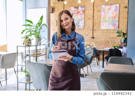 Young woman worker, owner in apron holding notepad pen in restaurant, coffee shop Young woman worker, owner in apron holding notepad pen in restaurant, coffee shop 119492342