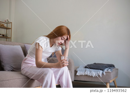 Woman Sitting on Sofa Holding Head in Pain, Suffering from Headache or Stress, Indoor Home Setting Woman Sitting on Sofa Holding Head in Pain, Suffering from Headache or Stress, Indoor Home Setting 119493562
