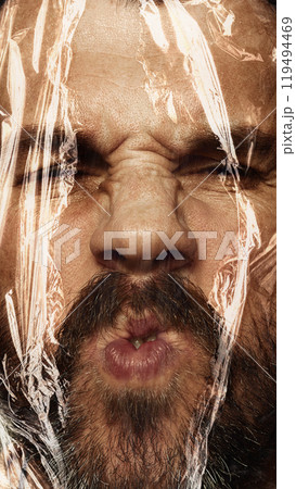 Close-up of male face wrapped in plastic cover. Man with moustaches expressing funny face with eyes closed. Conceptual photography. 119494469