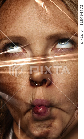 Close-up of female face wrapped in plastic cover. Woman grimacing, making funny face expressing boredom. Conceptual photography. 119494472