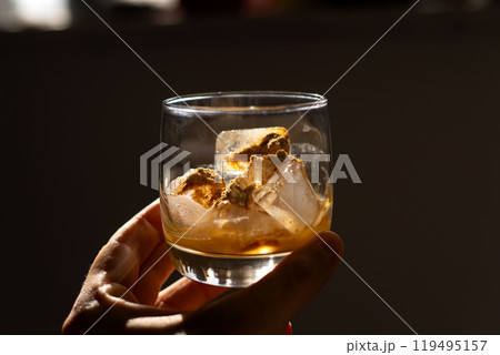 A glass filled with ice cubes and powdered Amanita muscaria mushroom on its surface, held in the hand, illuminated by natural sunlight. Preparing a homemade fly agaric elixir 119495157
