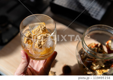 A glass filled with ice cubes and powdered Amanita muscaria mushroom on its surface, held in the hand, illuminated by natural sunlight. Preparing a homemade fly agaric elixir A glass filled with ice cubes and powdered Amanita muscaria mushroom on its surface, held in the hand, illuminated by natural sunlight. Preparing a homemade fly agaric elixir 119495158