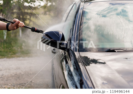 A person washing a black car with a pressure washer on a sunny day amidst greenery 119496144