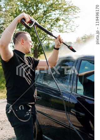 A man using a pressure washer to clean the side of a black car in a sunny outdoor setting A man using a pressure washer to clean the side of a black car in a sunny outdoor setting 119496170
