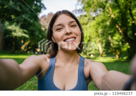Smiling pregnant woman sitting on grass and makes self-portrait on smartphone after workout 119498237