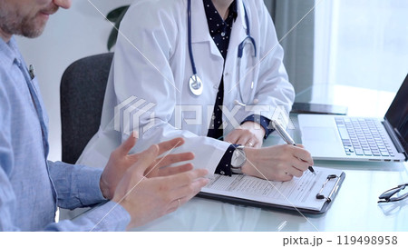 Doctor and patient are talking in clinic after usual health check up. Female physician is making notes awhile man wearing blue shirt is describing something. Medicine concept Doctor and patient are talking in clinic after usual health check up. Female physician is making notes awhile man wearing blue shirt is describing something. Medicine concept 119498958