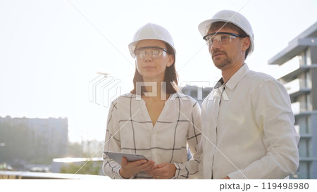 Two architects colleagues, wearing safety helmets and glasses, are standing near each other and discussing a construction site progress during sunrise or sunset. Architecture and engineering Two architects colleagues, wearing safety helmets and glasses, are standing near each other and discussing a construction site progress during sunrise or sunset. Architecture and engineering 119498980