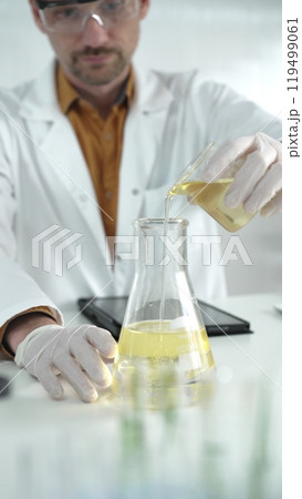 Man scientist with lab coat, white gloves, and protective glasses, is pouring a yellow oily liquid from one beaker to another near microscope in a laboratory, vertical close-up view Man scientist with lab coat, white gloves, and protective glasses, is pouring a yellow oily liquid from one beaker to another near microscope in a laboratory, vertical close-up view 119499061