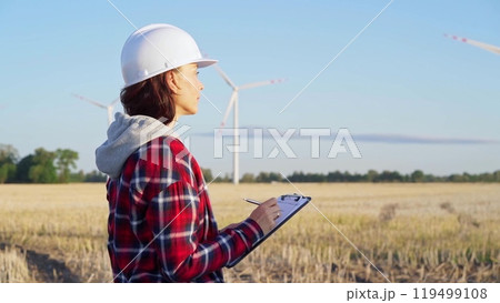 Female engineer wearing a white protective helmet is taking notes with a clipboard in a field with wind turbines, as the sun sets. Clean energy concept 119499108