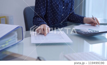 A female accountant with blue dotted blousy is using a laptop computer and calculator to calculate taxes at a glass desk in the office A female accountant with blue dotted blousy is using a laptop computer and calculator to calculate taxes at a glass desk in the office 119499190