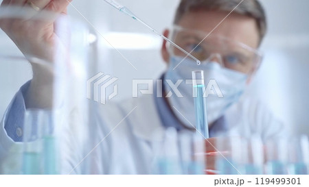 Man with safety glasses and blue mask is working with lab tubes using a pipette in a modern laboratory, close-up of researcher. Science and medicine Man with safety glasses and blue mask is working with lab tubes using a pipette in a modern laboratory, close-up of researcher. Science and medicine 119499301