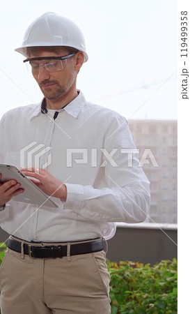 Male architect, wearing a white shirt and helmet, is using a digital tablet while inspecting a building site in foggy weather. Architecture and engineering concept 119499358