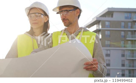 Architects or engineers wearing safety helmets and vests holding blueprint and discussing something on construction site at sunrise, front view. Architecture and engineering 119499402