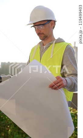 Man an architect or engineer wearing safety hard hat and vest holding blueprint and looking to construction site at sunrise, front vertical view. Architecture concept Man an architect or engineer wearing safety hard hat and vest holding blueprint and looking to construction site at sunrise, front vertical view. Architecture concept 119499403