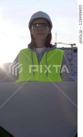 Female architect or engineer wearing safety hard hat and vest holding blueprint while inspecting a construction site at sunrise in early morning, front vertical view. Architecture concept 119499405