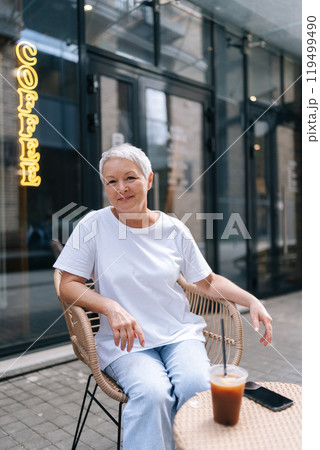 Charming elderly woman savoring iced coffee outside bustling cafe, appearing peaceful and at ease on sunny day, with smartphone close by to stay connected, smiling looking at camera. 119499490