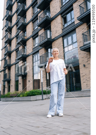 Vertical low-angle portrait of stylish senior woman drinking morning coffee outside modern city apartment, looking up with hope and contentment, enjoying break and savoring moment in retirement. 119499500