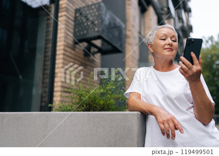 Portrait of elegant senior woman standing in urban setting, leaning on concrete wall and using smartphone, looking at phone screen with pleasant expression, enjoying free time in retirement 119499501
