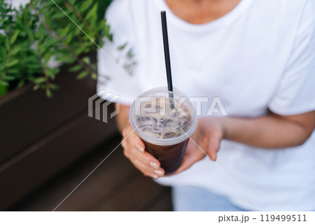 Close-up cropped shot of unrecognizable woman in white t-shirt holding takeaway plastic cup with cold coffee drink outdoors. Cup of iced bubble tea with straw closeup in female hand. 119499511