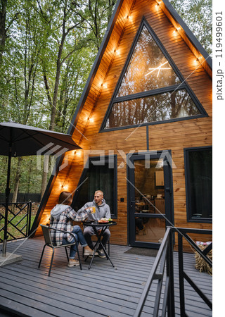 Couple enjoying drinks on a cozy wooden cabin terrace, surrounded by nature, relaxing getaway 119499601