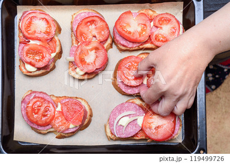 A woman's hand places a tomato slice on a sandwich before baking. 119499726