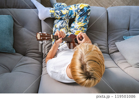 Teen boy plays ukulele guitar on modern couch. 119501086