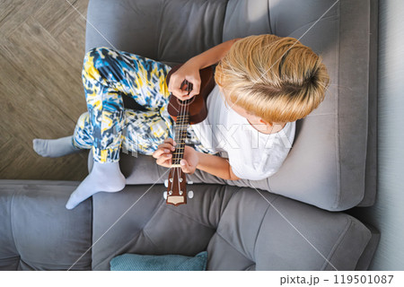 Teen boy plays ukulele guitar on modern couch. Teen boy plays ukulele guitar on modern couch. 119501087