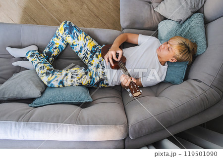 Young musician relaxing on sofa while strumming ukulele, surrounded by cushions. 119501090
