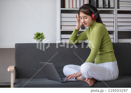 Young Woman Studying Online from Home on Laptop, Wearing Headphones, Sitting on Couch in Modern Living Room, Engaged in E-Learning, Remote Education, and Online Courses 119503297