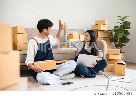 Happy Couple Running a Small Online Business from Home, Surrounded by Shipping Boxes, Working Together on a Laptop, Celebrating Success with a High Five 119503488