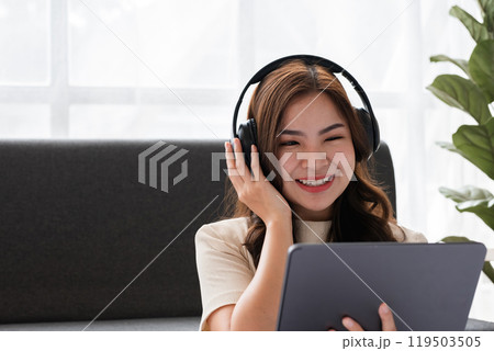 Young Girl Relaxing in Modern Living Room with Headphones and Tablet, Enjoying Music or Online Content, Bright and Cozy Home Interior with Natural Light and Green Plant in Background 119503505