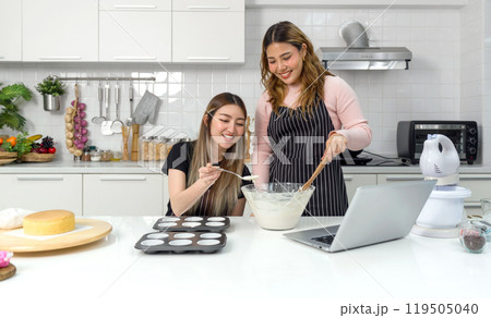 Two young women in an apron make dessert in the kitchen while looking at online recipe guide book on laptop computer. 119505040