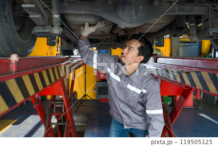Young mechanic in gray uniform maintenance or inspection work on a vehicle that is raised on a lift. Young mechanic in gray uniform maintenance or inspection work on a vehicle that is raised on a lift. 119505125