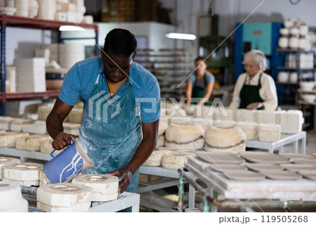 Slip casting - african american worker pours slip mass from jug into plaster mold in pottery factory 119505268