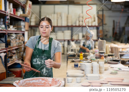 Young female worker in pottery factory lowers fired clay plates and cups into container with glaze Young female worker in pottery factory lowers fired clay plates and cups into container with glaze 119505270