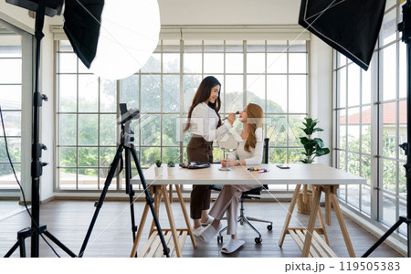 Two asian women creating content for social media. They are using a studio setup that include a camera mounted on a tripod, lighting equipment and various makeup product on the table. Two asian women creating content for social media. They are using a studio setup that include a camera mounted on a tripod, lighting equipment and various makeup product on the table. 119505383