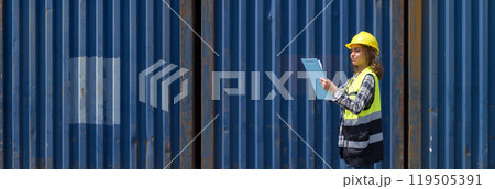 Young caucasian woman with safety vest and yellow hardhat checking shipping schedule on clipboard, planning for next shipment. A large cargo container is in the background 119505391