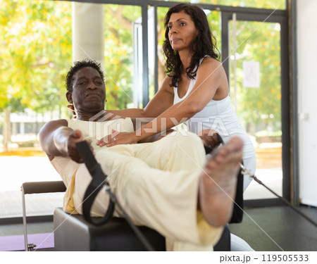 Focused hispanic instructor helping African American man practicing pilates. Individual coach Focused hispanic instructor helping African American man practicing pilates. Individual coach 119505533