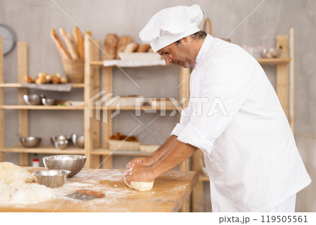 Male baker stands at work bench, kneading and shaping dough to make bread, croissants and baguettes 119505561
