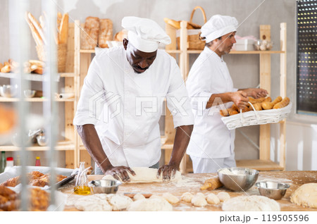 Skilled black baker rolling dough into long loaf shape Skilled black baker rolling dough into long loaf shape 119505596