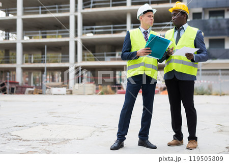 Two civil engineers are discussing the construction plan, holding documents and studying them Two civil engineers are discussing the construction plan, holding documents and studying them 119505809